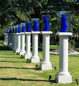 A row of Chilstone cast stone pedestals in a park displaying bright blue statues in glass cabinets for an art exhibition. 